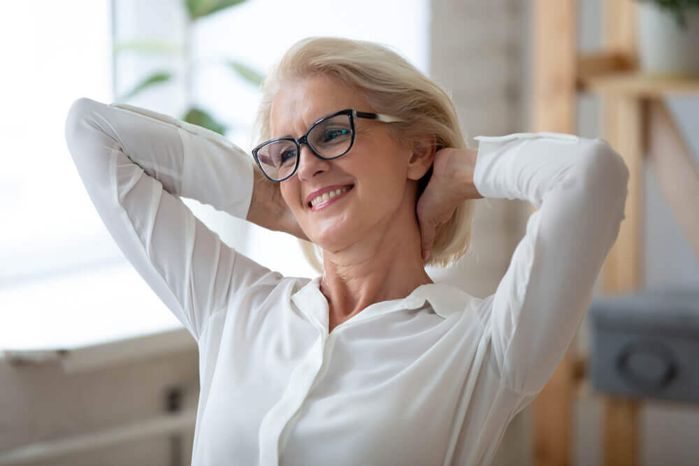 Elegant older woman with short blonde hair and black-rimmed glasses, smiling contentedly while stretching her arms behind her head. Perfect example of stylish glasses for women over 50, exuding confidence and comfort