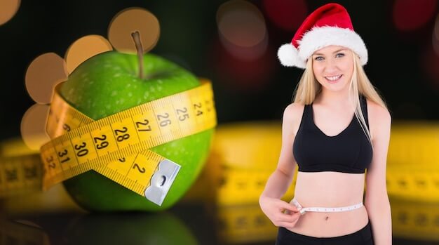 Young woman in a Santa hat measuring her waist with a tape measure, standing beside a green apple wrapped in a measuring tape, representing easy solutions for weight loss during the holiday season